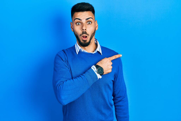 Joven hombre hispano con barba llevando suéter azul casual sorprendido señalando con el dedo hacia un lado, boca abierta expresión asombrada.  - Foto, imagen