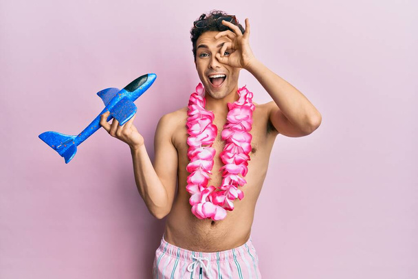 Fiatal, jóképű férfi fürdőruhát és Hawaii lei-t visel, kezében repülő játékkal, boldogan mosolyog, oké jel, kéz a kézben nézi az ujjait.  - Fotó, kép