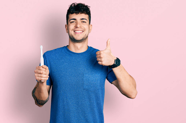 Giovane uomo ispanico in possesso di spazzolino da denti elettrico sorridente felice e positivo, pollice in su facendo eccellente e segno di approvazione  - Foto, immagini