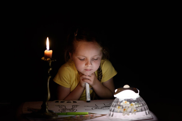 Mała dziewczynka rysuje postacie fantasy w pobliżu świecy w ciemnym pokoju. Dziecko robi ulubioną rzecz podczas przerwy w dostawie prądu. - Zdjęcie, obraz