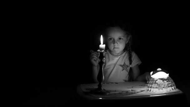 小さな女の子は暗い部屋でろうそくの近くにファンタジーキャラクターを描きます。停電時に好きなことをする子供。黒と白で. - 写真・画像