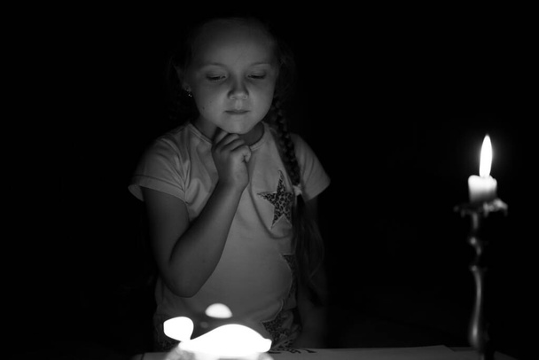 Little girl draws fantasy characters near candle in a dark room. Child doing favorite thing during a power outage. In black and white colors. - Photo, Image