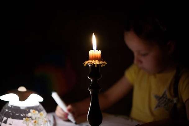 Το κοριτσάκι ζωγραφίζει φανταστικούς χαρακτήρες κοντά σε κεριά σε ένα σκοτεινό δωμάτιο. Παιδί κάνει το αγαπημένο πράγμα κατά τη διάρκεια μιας διακοπής ρεύματος. - Φωτογραφία, εικόνα