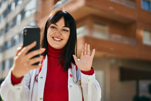 Nuori brunette nainen yllään lääkäri univormu tekee videocall älypuhelimella kaupungin - Valokuva, kuva