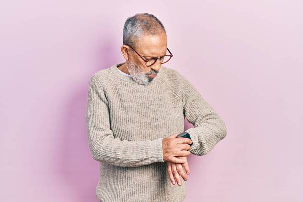 Hezký starší muž s vousy na sobě ležérní svetr a brýle kontrolu času na hodinkách na zápěstí, uvolněný a sebevědomý  - Fotografie, Obrázek