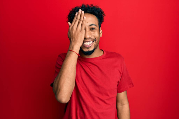 Молодой африканский американец с бородой в обычной красной футболке, прикрывающей один глаз рукой, уверенно улыбающийся на лице и удивляющий эмоции.  - Фото, изображение