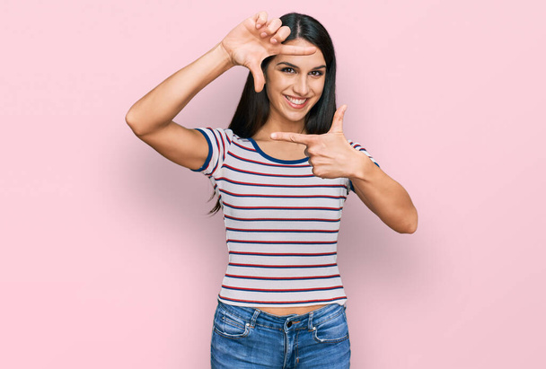 Junge hispanische Mädchen tragen lässig gestreiftes T-Shirt lächelnd Rahmen mit Händen und Fingern mit glücklichem Gesicht. Kreativität und Fotografiekonzept.  - Foto, Bild