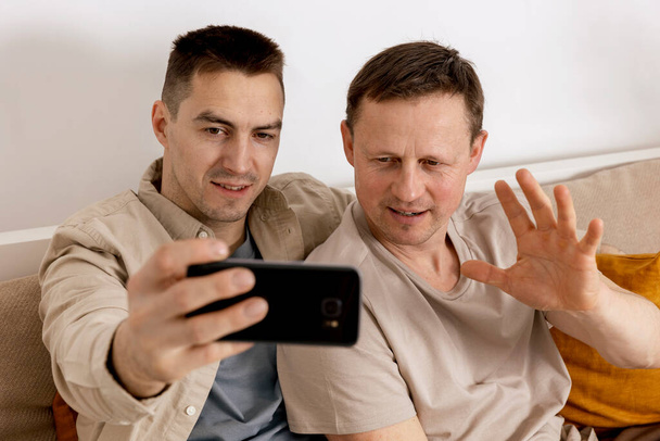 Šťastný gay pár s ležérní oblečení drží smartphone a mluvit s přáteli on-line, s videohovory. Homosexuální vztahy a alternativní láska. - Fotografie, Obrázek