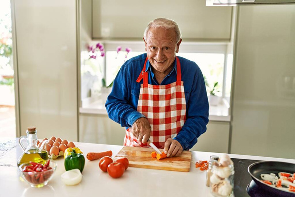Homem sênior sorrindo confiante cortando cenoura na cozinha - Foto, Imagem