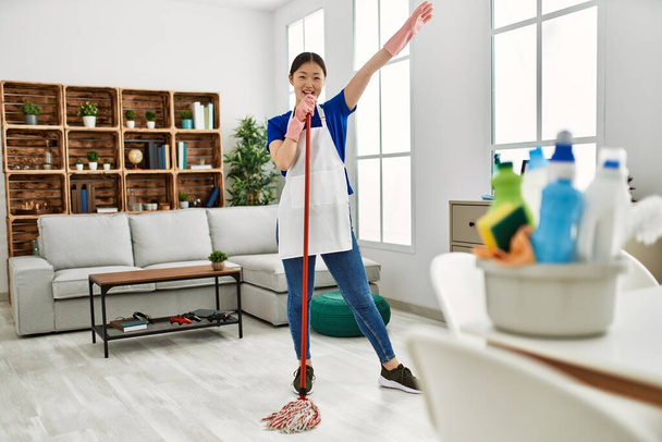 Young chinese housewife smiling happy singing and dancing using mop at home. - Photo, Image