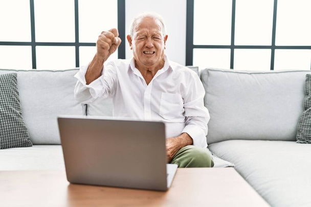 Senior man using laptop at home sitting on the sofa angry and mad raising fist frustrated and furious while shouting with anger. rage and aggressive concept.  - Foto, immagini