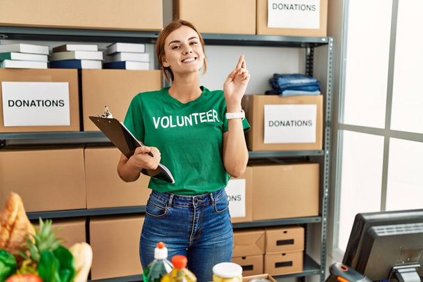 Młoda brunetka nosząca koszulę wolontariuszki na darowiznach stoi gestykulując trzymając kciuki uśmiechając się z nadzieją i zamkniętymi oczami. szczęście i przesądna koncepcja.  - Zdjęcie, obraz