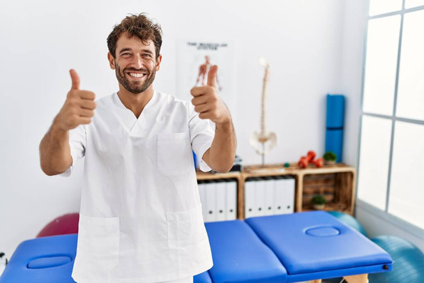 Young handsome physiotherapist man working at pain recovery clinic success sign doing positive gesture with hand, thumbs up smiling and happy. cheerful expression and winner gesture.  - 写真・画像