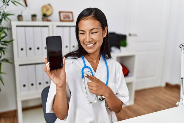 Jovem latina vestindo uniforme médico segurando smartphone na clínica - Foto, Imagem