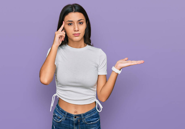 Jong Latijns-Amerikaans meisje draagt casual wit t-shirt verward en geïrriteerd met open palm met kopieerruimte en wijzende vinger naar het voorhoofd. Denk er eens over na..  - Foto, afbeelding