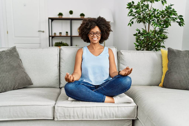 Young african american woman doing yoga sitting on sofa at home - Foto, imagen