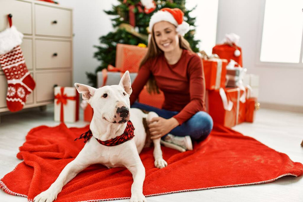 Молодая белая женщина, уверенно улыбающаяся, сидя дома с собакой у рождественской елки - Фото, изображение