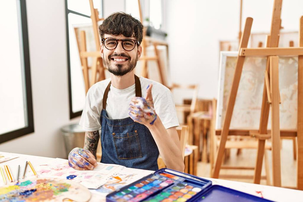 Homme hispanique avec barbe au studio d'art faisant des pouces heureux geste avec la main. approuvant l'expression regardant la caméra montrant le succès.  - Photo, image