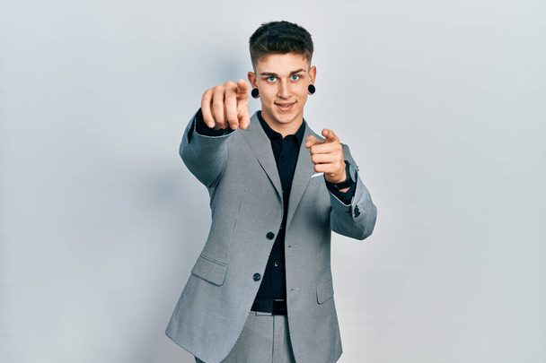 Jeune garçon caucasien avec dilatation des oreilles portant une veste d'affaires pointant vers vous et la caméra avec les doigts, souriant positif et joyeux  - Photo, image