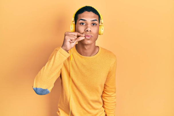 Jeune homme afro-américain écoutant de la musique à l'aide d'écouteurs bouche et lèvres fermées comme zip avec les doigts. secret et silencieux, tabou parlant  - Photo, image