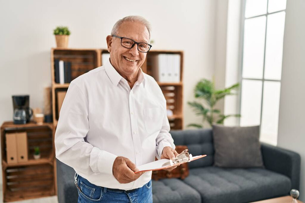 Hombre mayor psicólogo sonriendo confiado sujetando portapapeles en el centro de psicología - Foto, imagen