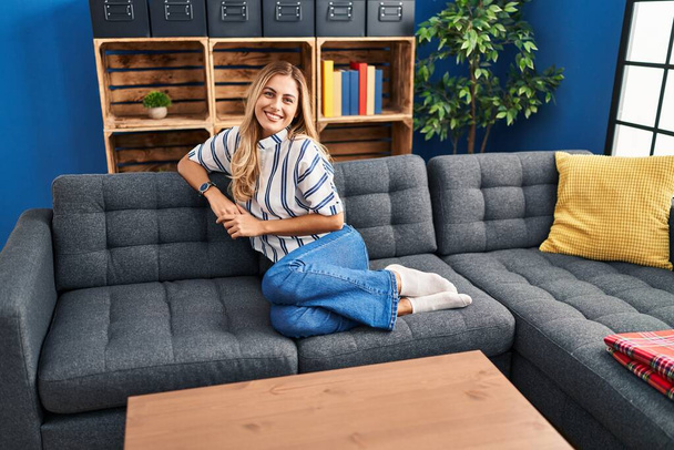 Молодая блондинка улыбается уверенно сидя дома на диване - Фото, изображение