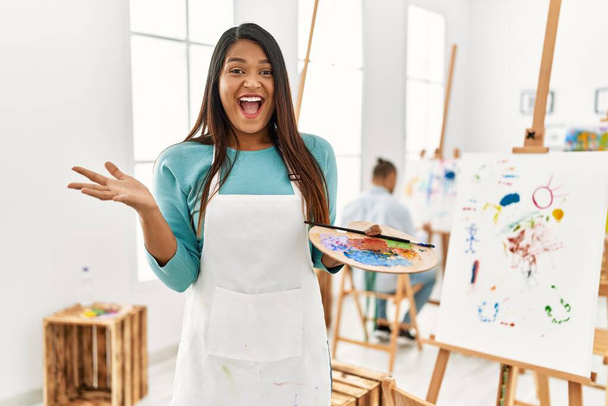 Young latin woman standing at art studio celebrating victory with happy smile and winner expression with raised hands  - Photo, Image