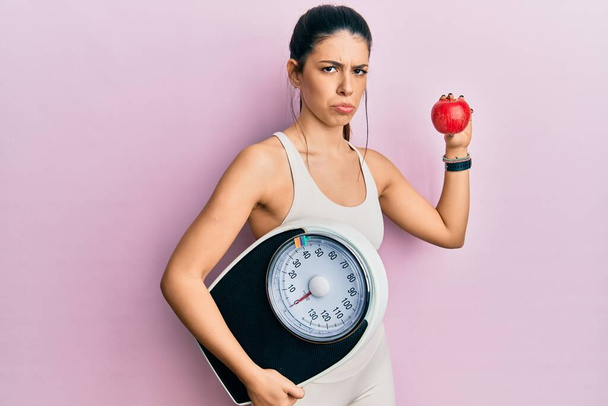 Fiatal spanyol nő visel sportruházat gazdaság súlymérő gép és alma szkeptikus és ideges, homlokráncolás ideges, mert a probléma. negatív személy.  - Fotó, kép