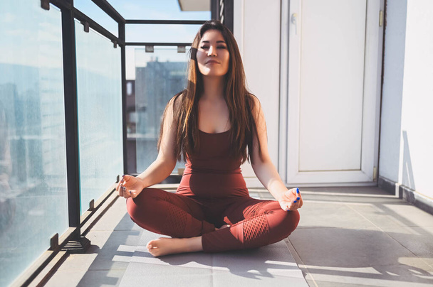 Beautiful Asian smiling fit woman doing yoga on sunny balcony in sport suit. Young happy lady on terrace at summertime. Big city view. Sports exercises and gymnastics, healthy way of life concept. - Photo, image