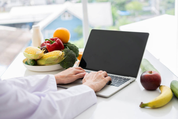 Portrait of Asian smiling female nutritionist typing on laptop computer for balanced corrective diet plan for patient or weight loss advice. Concept of healthy lifestyle, healthy food, dieting - Photo, Image