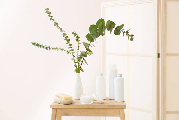 Different cosmetic products and vase with eucalyptus branches on wooden table in room - Photo, image