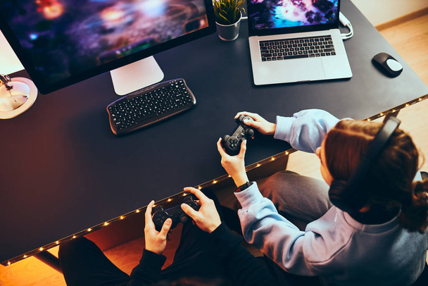 Friends playing video game at home. Gamers holding gamepads sitting at front of screen. Streamers girl and boy playing online in dark room lit by neon lights. Competition and having fun - Foto, Imagen