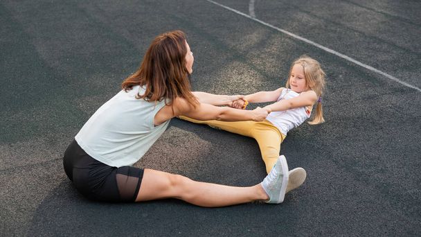 Mother and daughter go in for sports outdoors. Caucasian woman and little girl are engaged in fitness at the stadium. - 写真・画像