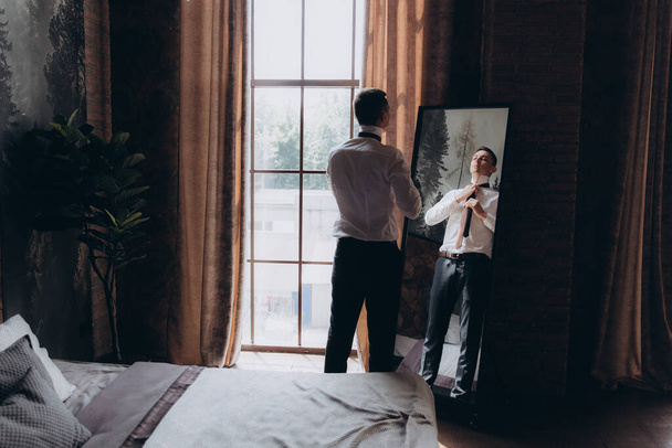 Morning of a stylish groom, man, in a black suit and tie, his preparations at the hotel, in a photo studio, fastening buttons and jacket, portraits - Fotó, kép