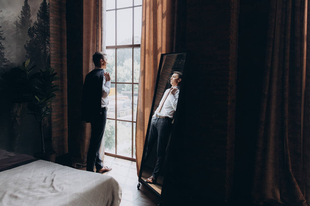 Morgens ein stilvoller Bräutigam, Mann, in schwarzem Anzug und Krawatte, seine Vorbereitungen im Hotel, im Fotostudio, Knöpfe und Jacke befestigen, Porträts - Foto, Bild