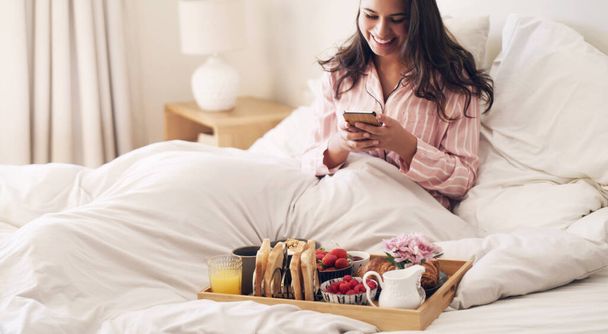 Paras tapa aloittaa päiväsi. Laukaus kaunis nuori nainen käyttää älypuhelinta, kun aamiainen sängyssä kotona. - Valokuva, kuva