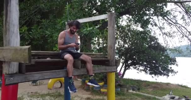 mladý muž pomocí smartphone ve dne se zeleným parkem v pozadí - Záběry, video