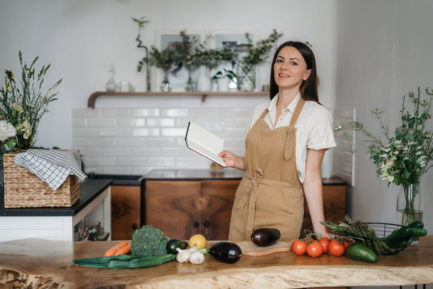 красивая молодая брюнетка женщина готовит на кухне по рецепту из ноутбука здоровое питание из свежих органических овощей смотрит в камеру - Фото, изображение