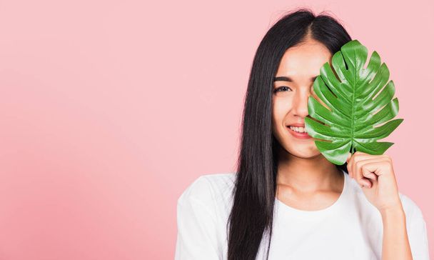 Beauty face. Portrait of Asian beautiful young woman with fresh healthy skin hold green monstera leaf on her face, Tropical Leaves, studio shot isolated on pink background, Skin body care spa concept - Foto, Imagem