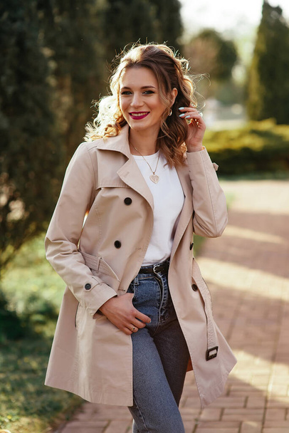 Pretty young woman with an up-do wearing a light grey trench coat, blue jeans, navy long knitted scarf and a leather messenger bag. - Valokuva, kuva