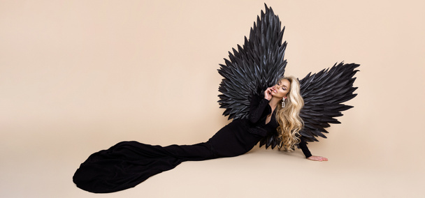 エレガントなファッション。エレガントな長い黒のドレスとスタジオでベージュの背景に大きな翼で見事なブロンドの女性。豪華な夜のファッション。グラマーファッションモデル。優しい女だな。翼を持つ女性。天使. - 写真・画像
