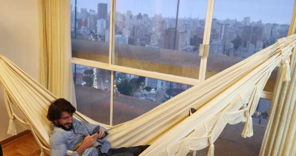 młody człowiek za pomocą smartfona leżącego w hamaku w domu - Materiał filmowy, wideo