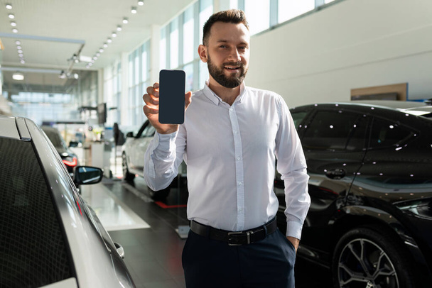 un vendedor en un concesionario de automóviles demuestra la pantalla de un teléfono inteligente en el fondo de los coches nuevos - Foto, Imagen