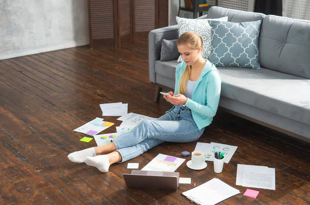 Young woman works with documents using smartphone and laptop while sitting on the floor at home. Student, entrepreneur or freelancer girl working or studying remotely via internet. - Foto, immagini