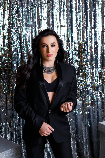 Portrait of a young beautiful woman with dark hair of European appearance. Dressed in a black jacket. Silver shiny background - Foto, Imagem