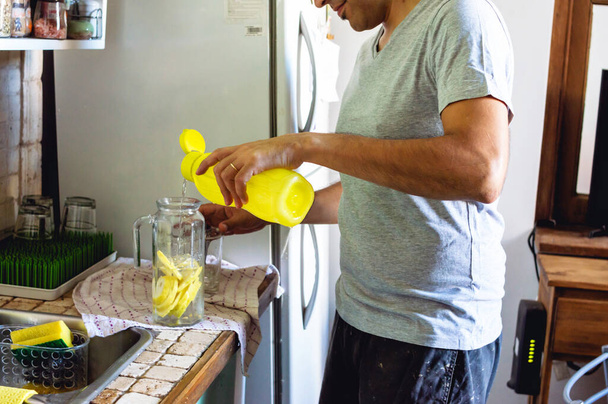 young man standing in the kitchen at home pouring water in a glass jug with lemon slices, lifestyle concept. - Foto, immagini