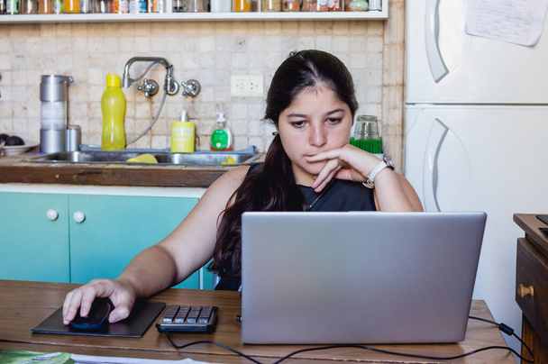 joven mujer caucásica trabajando en casa con su portátil y con una cara concentrada con una mano en el ratón y la otra mano tocando su labio, mirando a la pantalla de la computadora - Foto, Imagen
