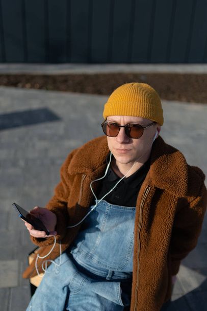 Απλή hipster κάθεται σε ένα παγκάκι στην πόλη, χρησιμοποιώντας smartphone και ακουστικά του. Υψηλής ποιότητας φωτογραφία - Φωτογραφία, εικόνα