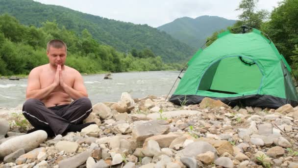 Le gros pratique le yoga près d'une rivière de montagne, suivi d'une tente verte. - Séquence, vidéo
