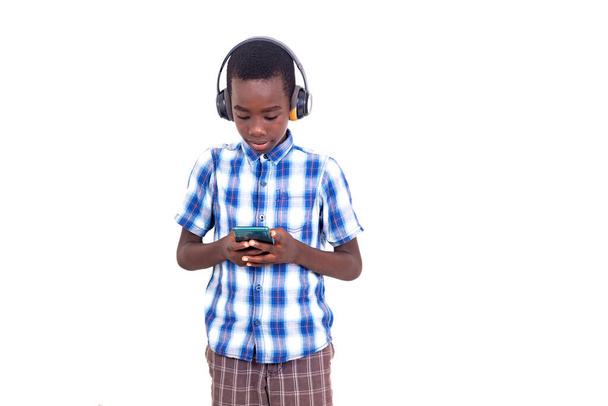 молодий хлопчик у картатій сорочці, що стоїть на білому тлі, спірально маніпулює мобільним телефоном, носячи гарнітури
. - Фото, зображення